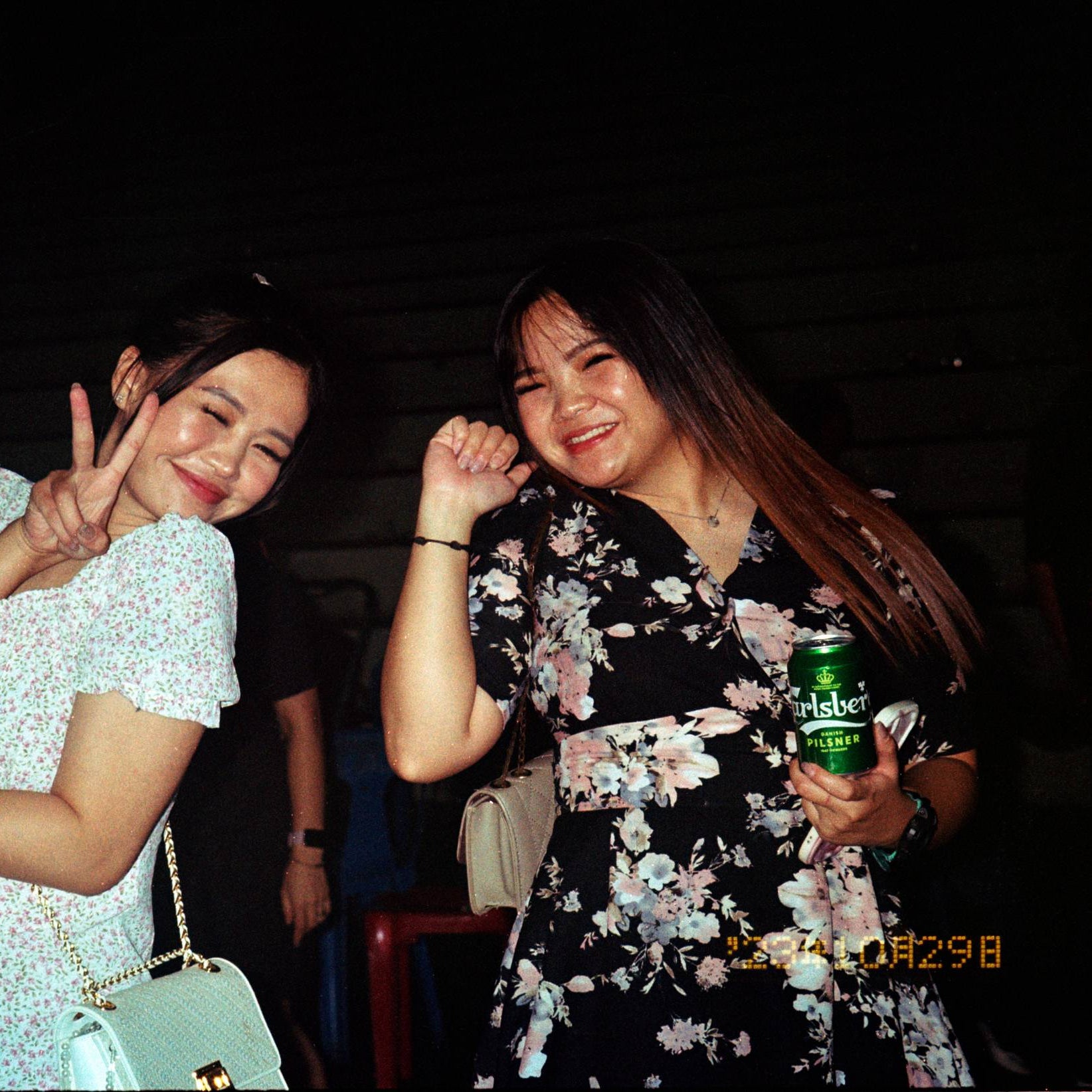 Two women posing together in a dark, social setting. The woman on the left wears a white floral short-sleeve dress and holds a light blue handbag, while the woman on the right wears a black floral dress and holds a green can of beer.