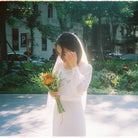 Woman in a white dress holding flowers in an outdoor setting with trees and buildings in the background.