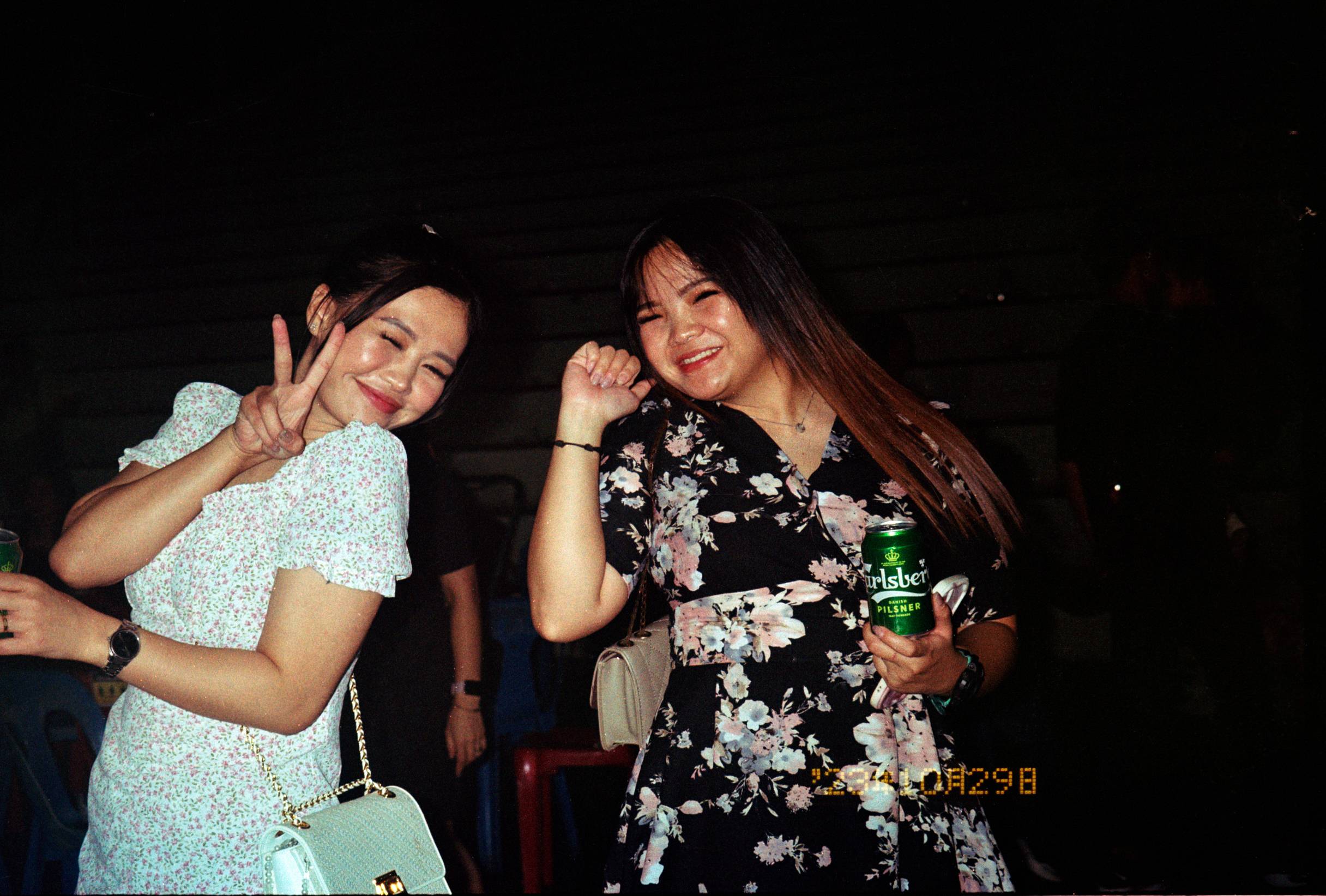 Two women posing together in a dark, social setting. The woman on the left wears a white floral short-sleeve dress and holds a light blue handbag, while the woman on the right wears a black floral dress and holds a green can of beer.