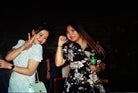 Two women posing together in a dark, social setting. The woman on the left wears a white floral short-sleeve dress and holds a light blue handbag, while the woman on the right wears a black floral dress and holds a green can of beer.