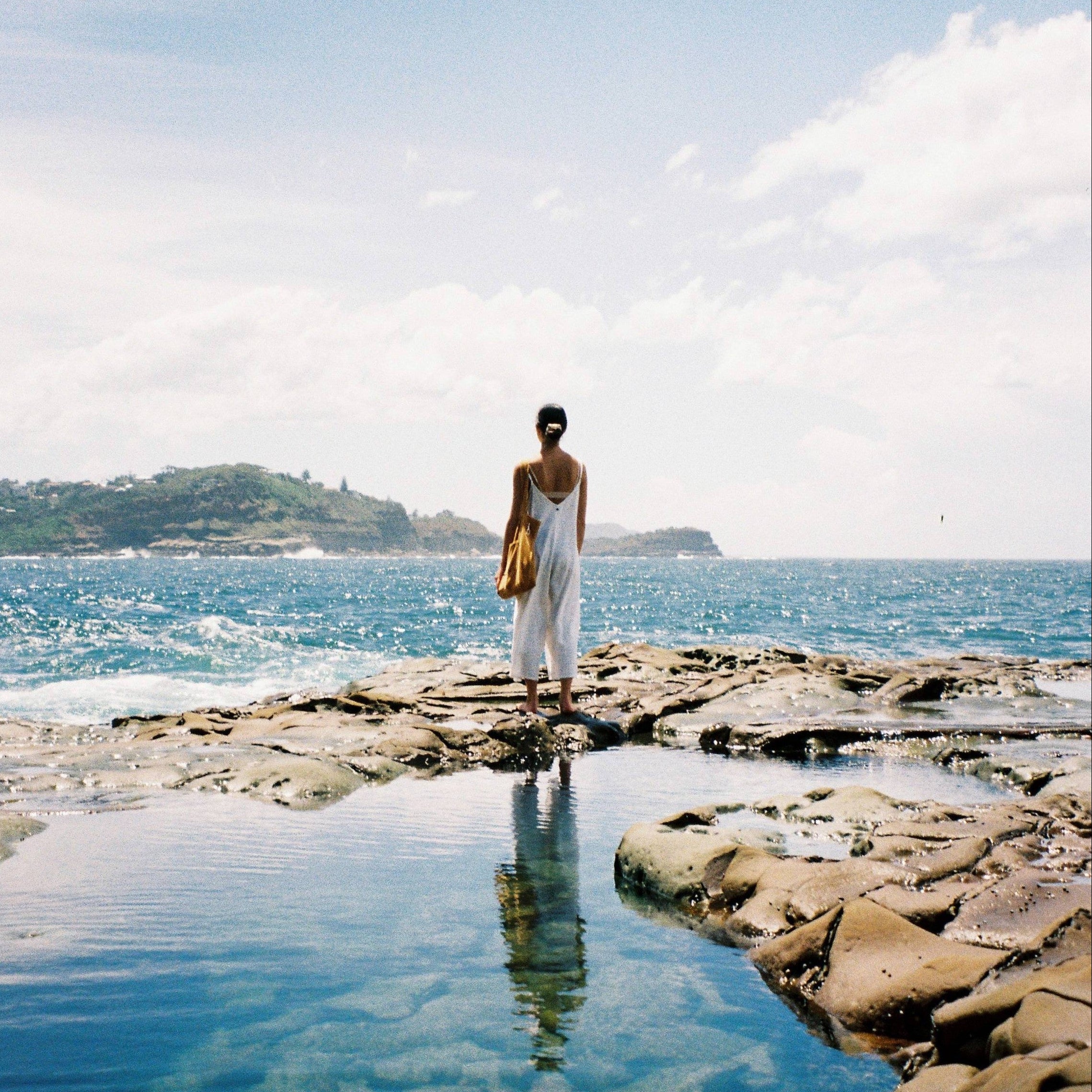 Person standing on a rocky outcrop by the ocean with clear blue water and sky.