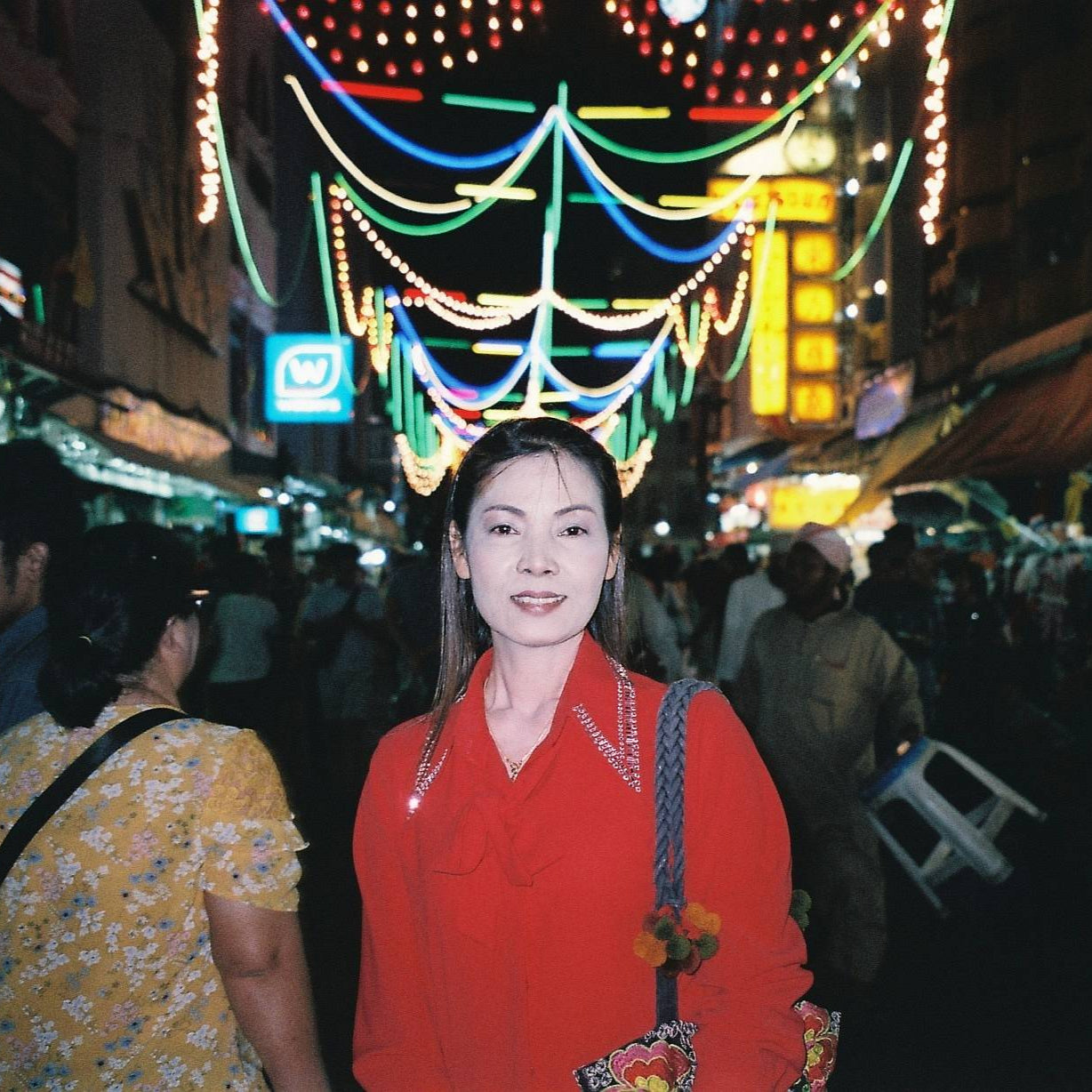 Woman in a red shirt standing in a bustling street at night with colorful lights and people around.