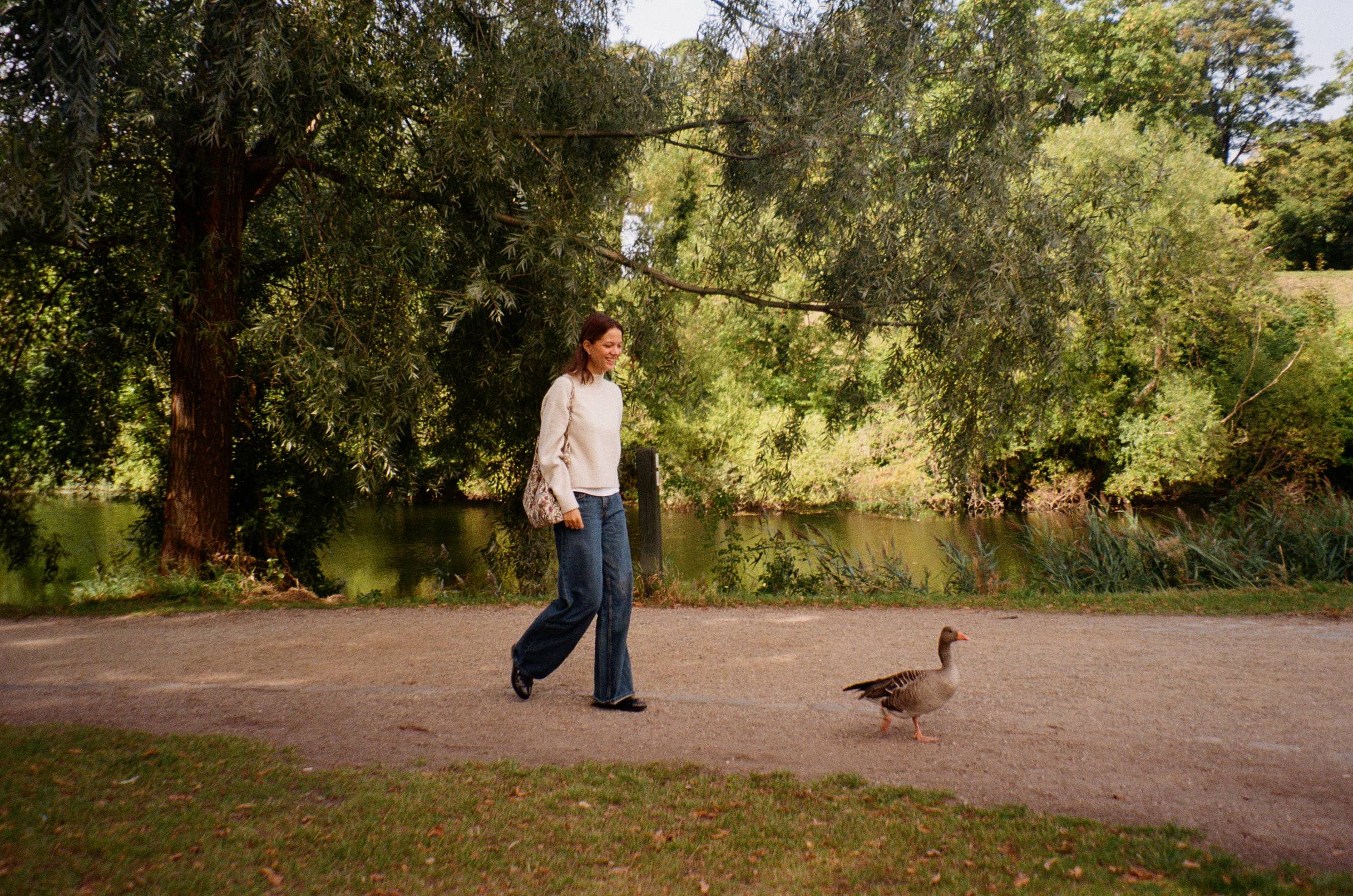 Woman walking with a duck on a path in a park with trees and water in the background