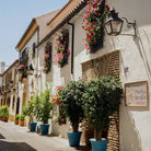 Street scene with white buildings, hanging flowers, and potted plants in a Mediterranean-style town.